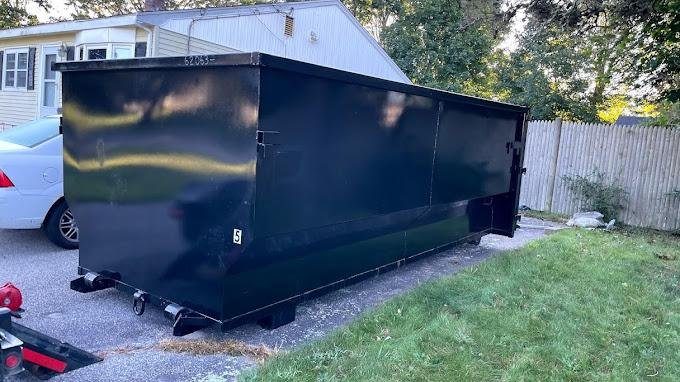 Black roll-off dumpster set at the edge of a lawn beside a paved driveway
