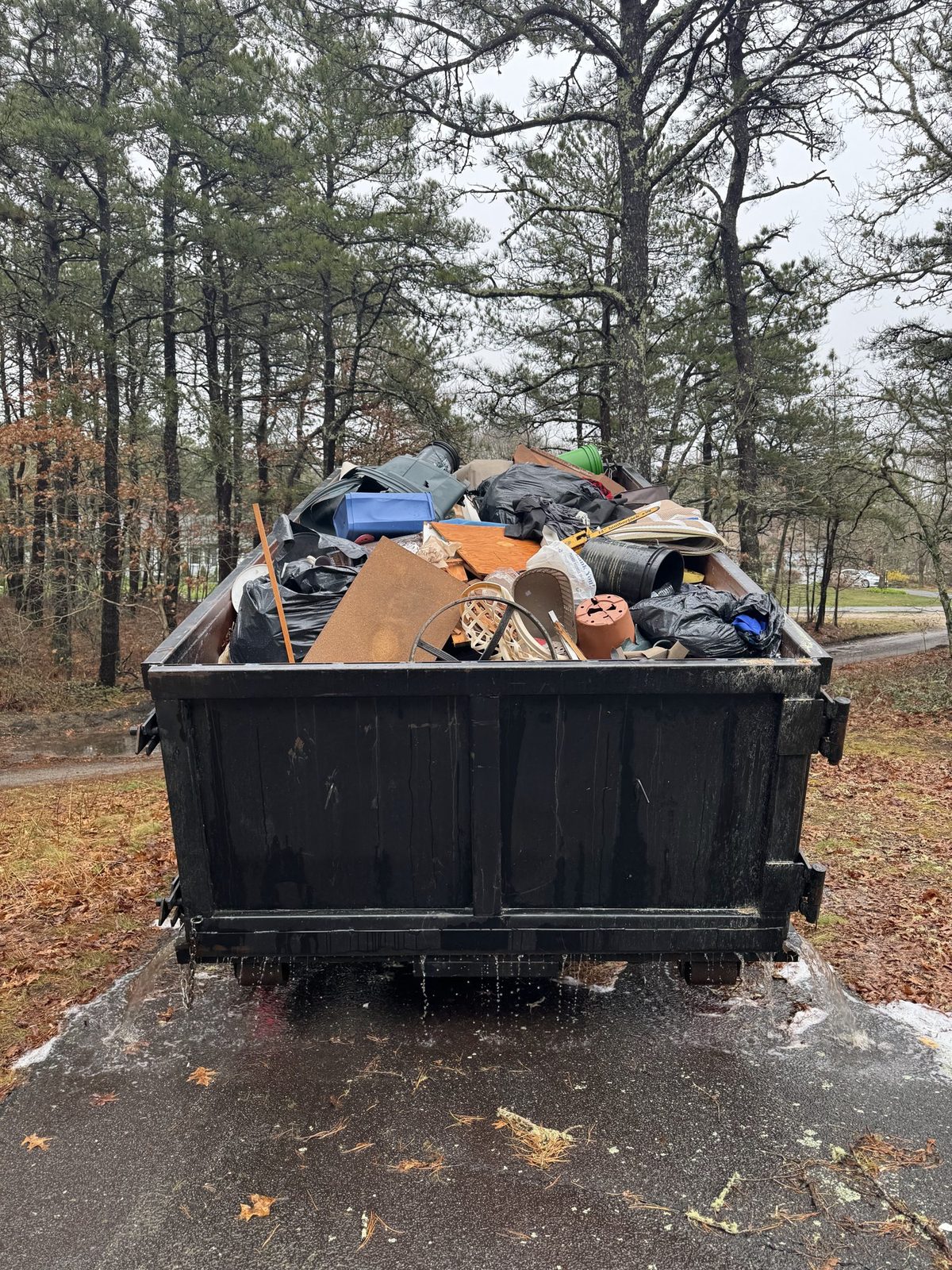 Roll-off dumpster filled with household cleanout items, boards and trash bags in a wooded yard