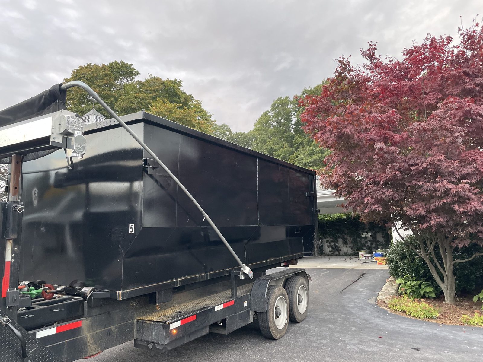 Tall black roll-off dumpster on a trailer in a driveway beside a red Japanese maple