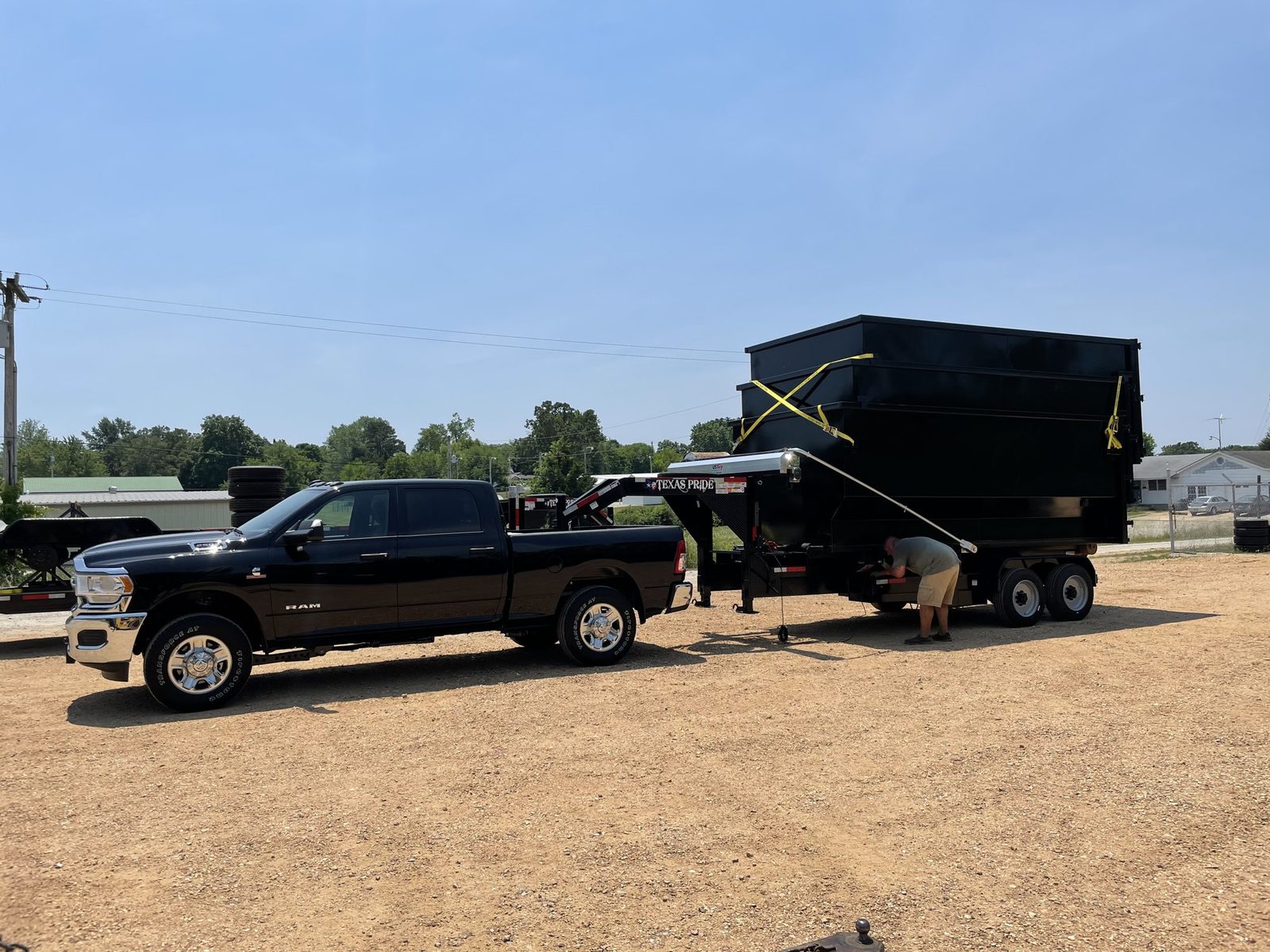 Pickup truck hitched to a gooseneck trailer carrying a tall black dumpster on a dirt lot
