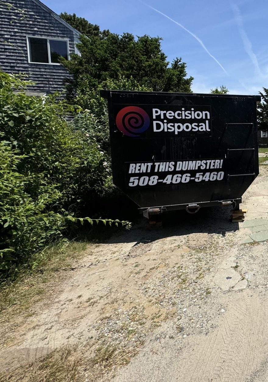 Precision Disposal roll-off dumpster with a "Rent This Dumpster! 508-466-5460" decal in a sandy driveway beside a shingled cottage