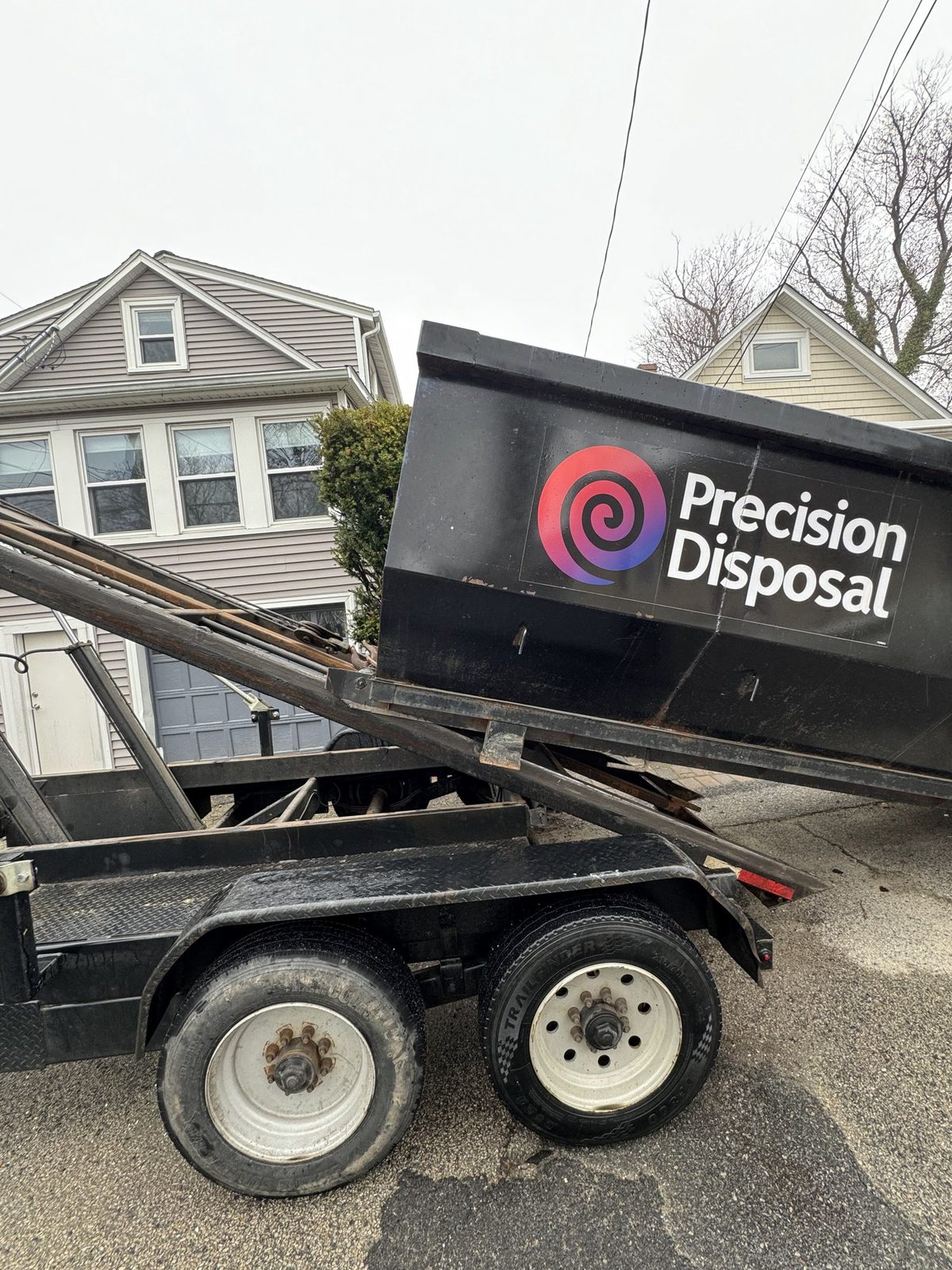 Precision Disposal dumpster tilted on its trailer in front of a two-story house