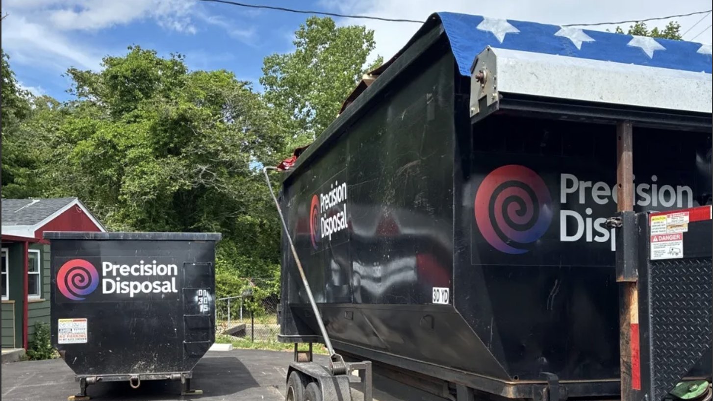 Precision Disposal roll-off dumpsters and a loaded dumpster trailer parked on a gravel lot