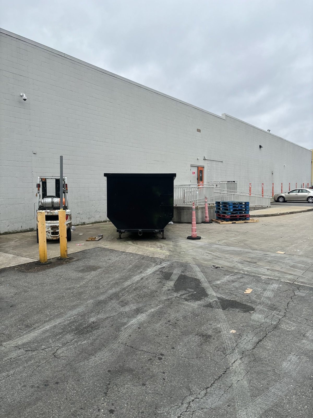 Black roll-off dumpster set beside a warehouse loading ramp, with safety cones and stacked pallets