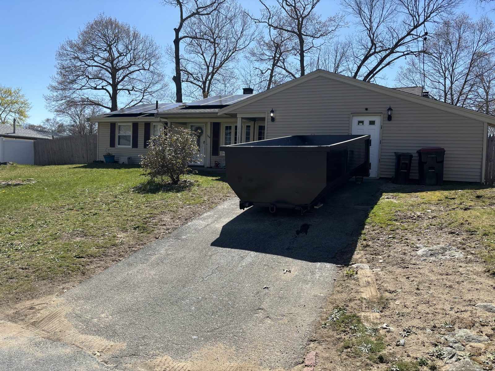 Black roll-off dumpster set in the driveway of a single-story ranch house, beside two trash bins