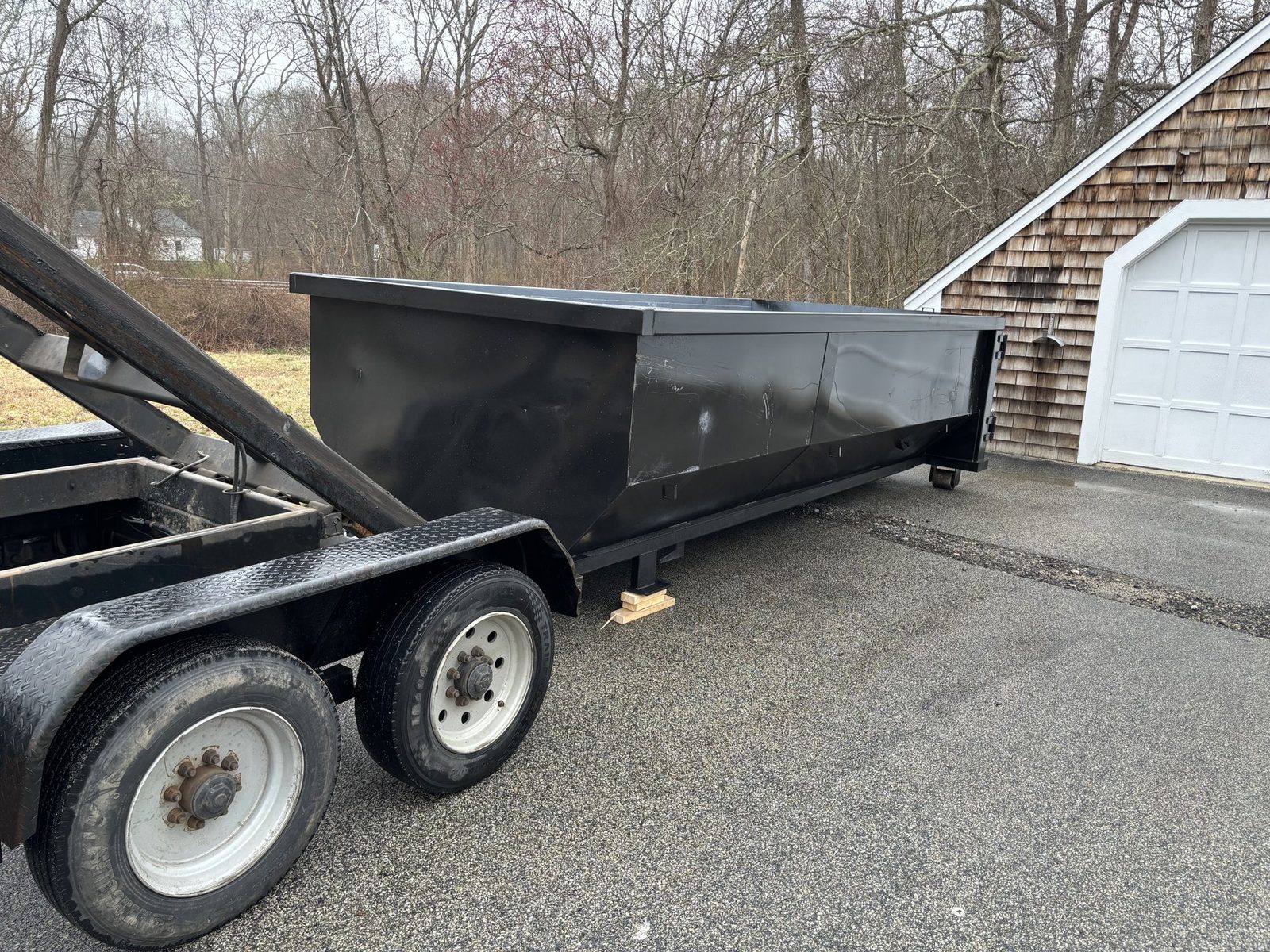 Black roll-off dumpster on a tilt trailer in a paved driveway next to a shingled garage
