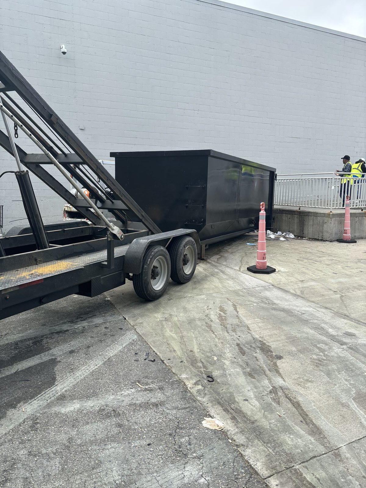 Roll-off dumpster on a tilt trailer beside a gray block commercial building, with an orange safety cone