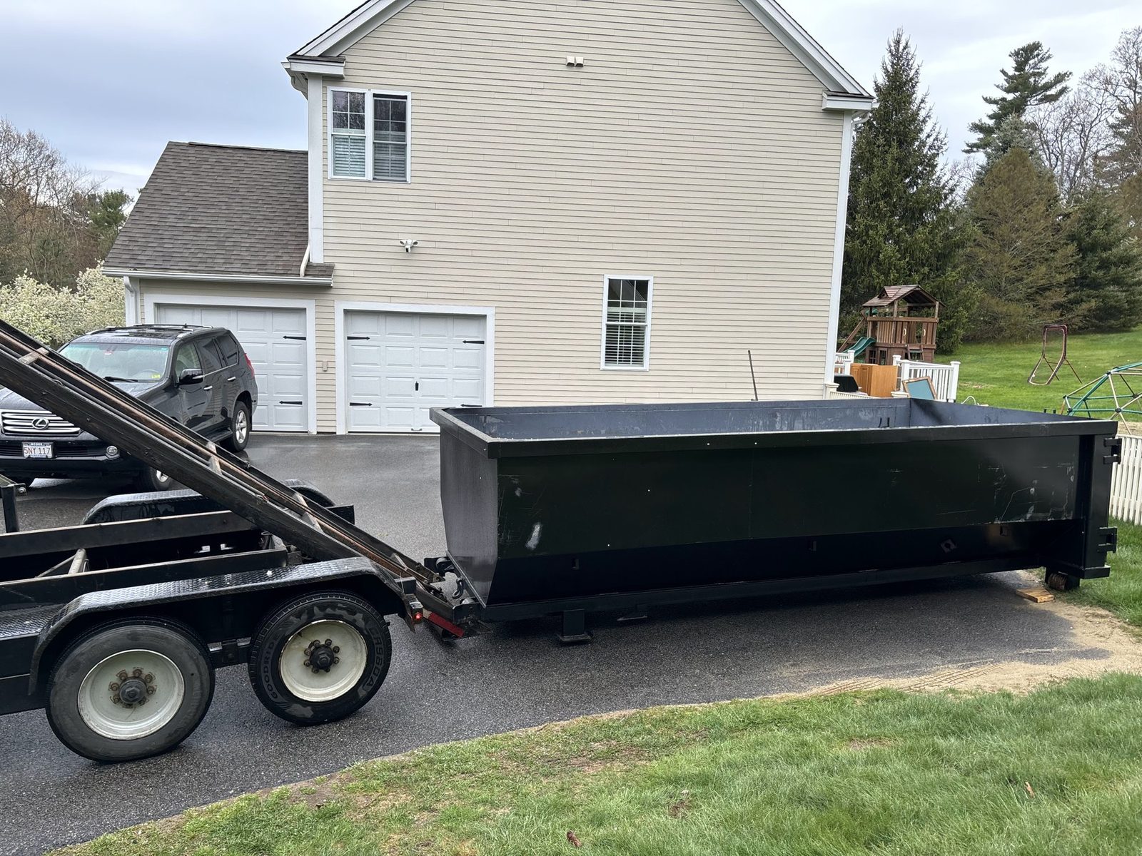 Roll-off dumpster sliding off a tilt trailer onto a paved driveway in front of a two-story house with a two-car garage